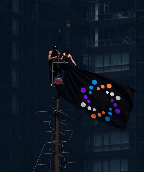The user wants a caption for the image based on the provided text.

1.  **Analyze the image:**
    *   Two people are on a high platform (looks like a crane or antenna structure) on top of a building.
    *   They are holding a large black flag.
    *   The flag has a logo on it: a circle made of dots in various colors (blue, orange, white, purple).
    *   The background is a dark, tall building with many windows.

2.  **Analyze the text:**
    *   "The problem with crypto trading in 2026 isn’t access to information, it’s that information is everywhere and useful signal is nowhere, that’s the exact problem @nodiens is built to solve, Nodiens is a unified crypto market intelligence platform that gives traders full visibili" (text cuts off but implies "visibility").
    *   Key entity: "@nodiens" and "Nodi