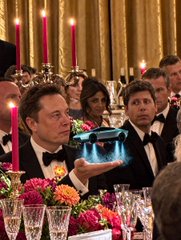 A photo of Elon Musk holding up a Grok-branded robot at a formal dinner event, with another man in a tuxedo sitting next to him amid elegant table settings.