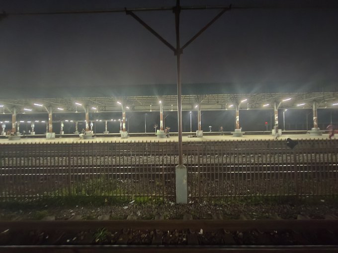 A nighttime view of a brightly lit, empty train station platform with multiple illuminated overhead canopies, viewed from across railroad tracks behind a metal fence.