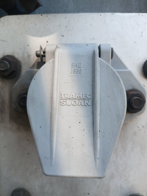 A close-up of a metallic T-bolt hose clamp branded 'TRAMEC SLOAN' with 'SAE J560' marking, bolted to a white surface.