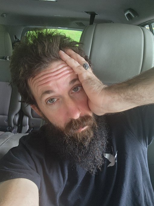 A selfie of a bearded man sitting inside a vehicle, resting his hand on his forehead with a tired or exasperated expression.