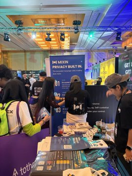 People browse materials at an MXDN 'IMPACTFUL IN' booth at what appears to be a tech convention or trade show, with banners and promotional items visible on the table.