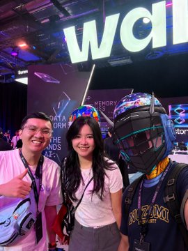 Three people pose for a photo at what appears to be a convention or exhibition, with a person wearing an elaborate black helmet and costume standing on the right.
