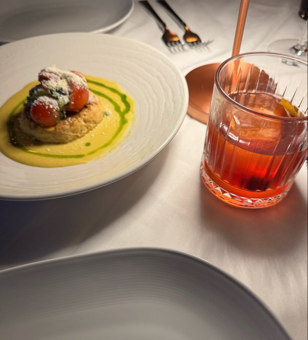 Elegant table setting with a white plate holding an appetizer of tomatoes and herbs over a yellow sauce with green oil drizzle, alongside a glass of amber-colored beverage.