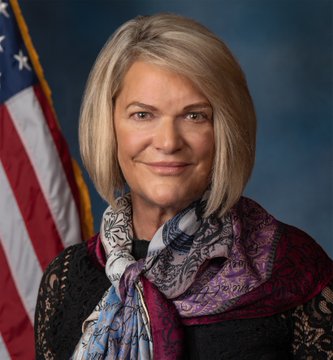 A professional portrait shows a woman with shoulder-length blonde hair wearing a black lace top and a vibrant purple and blue patterned scarf, posed in front of an American flag. The image accompanies news about Senator Cynthia Lummis's criticism that Democrats are holding the Crypto Clarity Act "hostage."