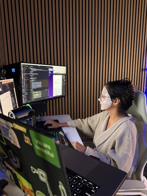 A woman wearing a sheet mask and a cozy robe sits at a dual-monitor crypto trading setup, with a Shure microphone and sticker-covered laptop in front of her, blending self-care with screen time. The setup reflects the post's theme of balancing birthday relaxation with the nonstop nature of crypto trading.