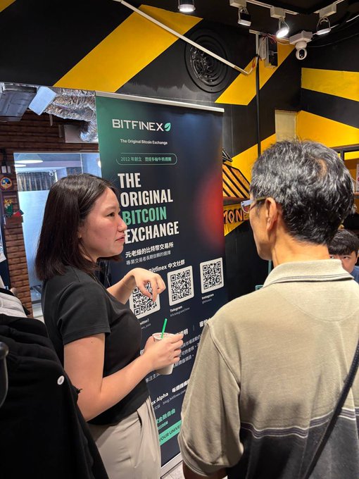 A Bitfinex promotional booth at what appears to be a convention or event, with a woman explaining the Bitcoin exchange services to two men, featuring Chinese text and QR codes.