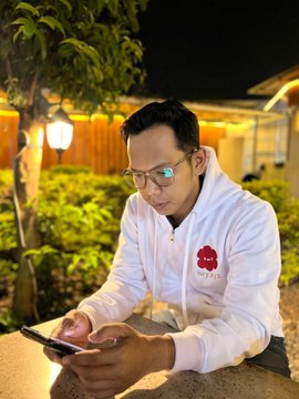 A man wearing glasses and a white zip-up jacket with a red emblem sits outdoors at night, looking down at his smartphone.