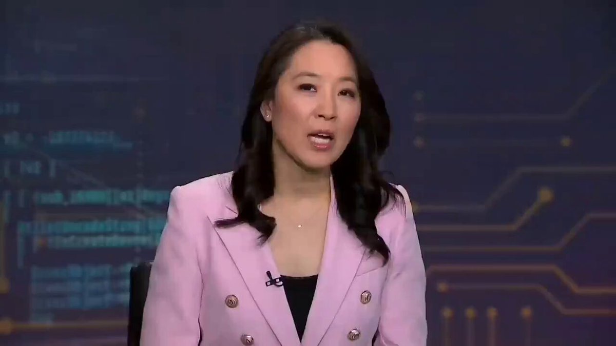 The image features a woman wearing a pink blazer, who is sitting behind a desk with her hands placed in front of her. She appears engaged and focused while looking directly into the camera during an interview-style conversation. The backdrop has an intriguing graphic design that includes abstract shapes and lines, adding visual interest to the setting. The text within the image refers to a recent market event or trend where Mastercard is contemplating investing in a cryptocurrency company worth $1.8B, which is XRP specifically, as it aims to integrate digital assets into its network.