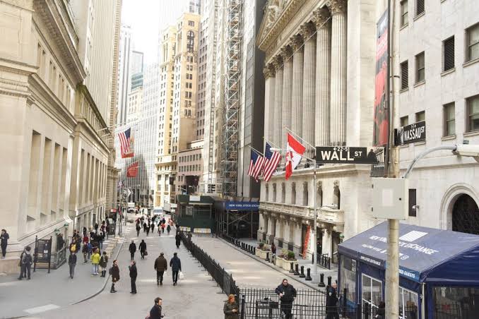 A street view of Wall Street in New York City, featuring the New York Stock Exchange with American flags and a 'Wall St' street sign visible.