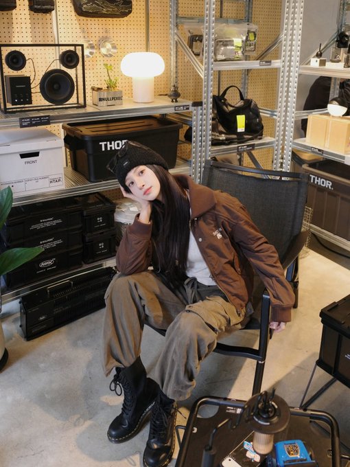 A young woman wearing a black beanie, brown jacket, and cargo pants sits on a chair in a room with shelves of storage boxes and audio equipment, including speakers labeled 'SMALLPERS'.