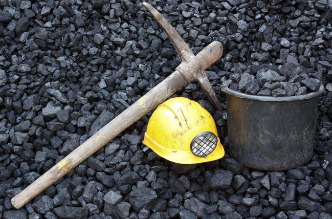 A yellow hard hat with a lamp, a pickaxe, and a metal bucket filled with coal arranged on a bed of black coal pieces.