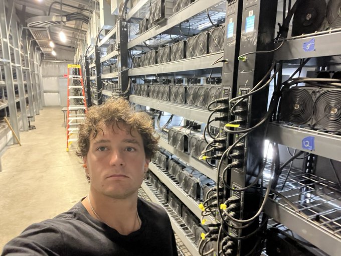 A young man with curly brown hair takes a selfie in what appears to be a Bitcoin mining facility, surrounded by racks of computer hardware and cooling fans.