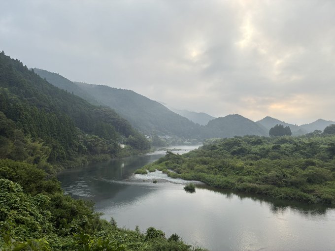 A serene landscape of a winding river flowing through lush green forested mountains under an overcast sky with the sun faintly breaking through the clouds.