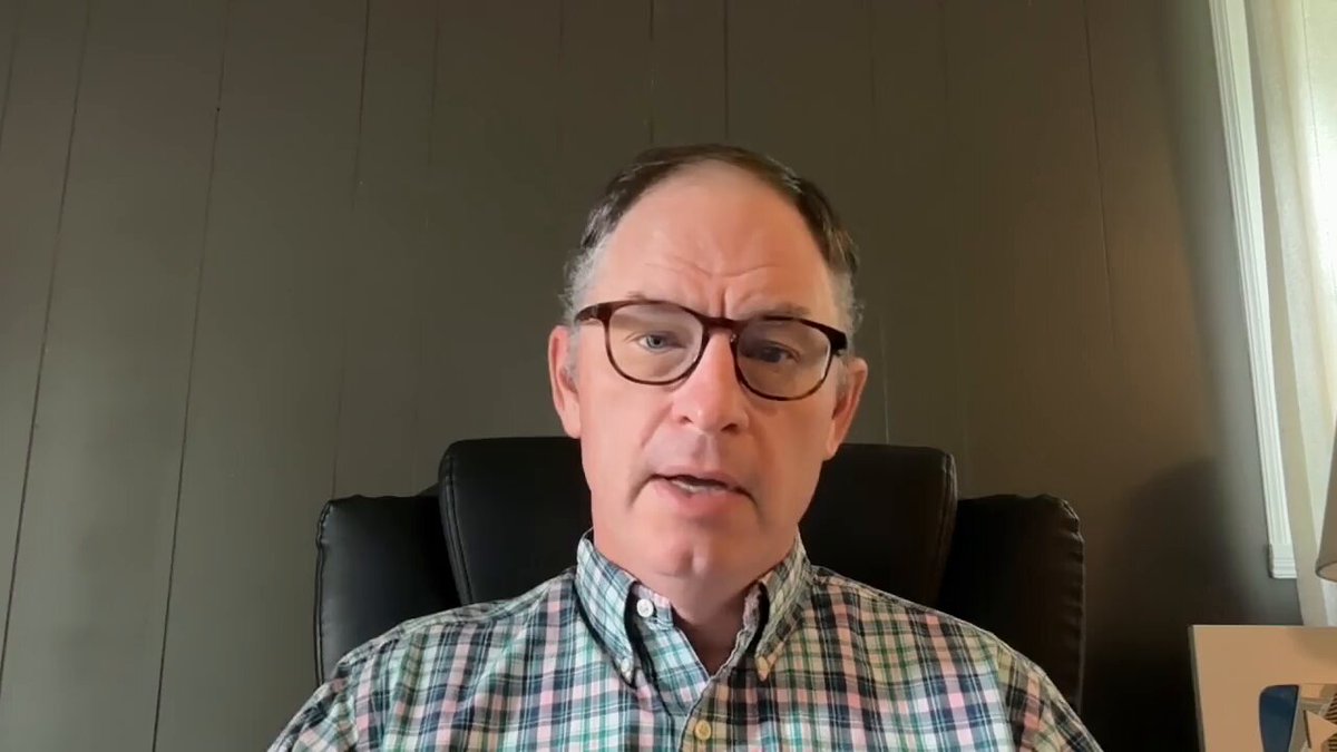 The image shows a man wearing glasses sitting in an office chair, looking at the camera with a serious expression. He appears to be engaged in a conversation or presentation. The background of the image features a gray wall, which contrasts with the black leather chair he is seated on. There are also two windows visible in the background, suggesting that the office space might have natural light coming through them.