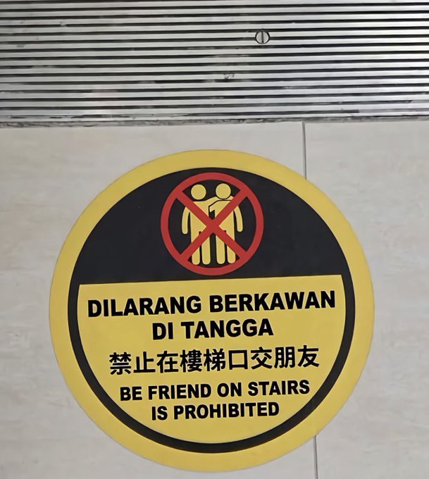 A yellow and black prohibition sign that reads 'DILARANG BERKAWAN DI TANGGA' (Malay), '禁止在楼梯口交朋友' (Chinese), and 'BE FRIEND ON STAIRS IS PROHIBITED' (English), featuring a graphic of two people with a red prohibition symbol over them.
