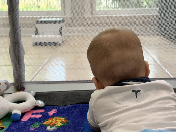 A baby lying on its stomach on a colorful play mat, looking out a glass door toward a white pet feeder on the patio.