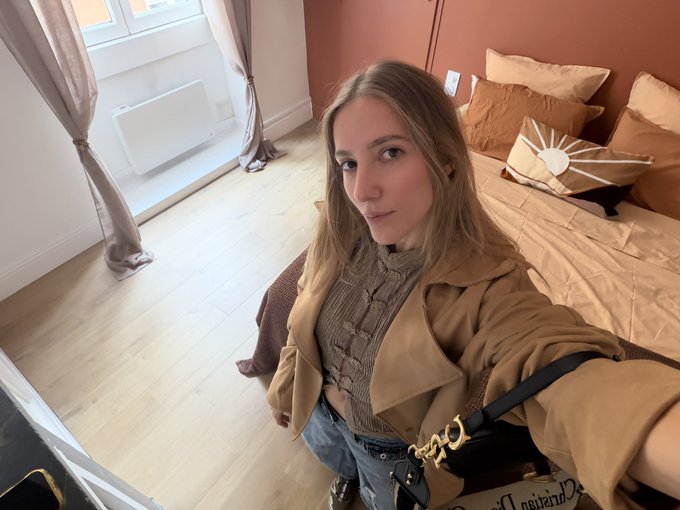 A selfie of a young woman with long blonde hair wearing a tan trench coat over a brown top, standing in a bedroom with peach-colored walls and bedding.