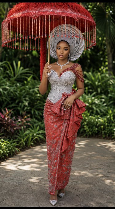 A woman wearing a traditional red and silver Nigerian bridal gown and silver head tie, holding a large ornate red parasol with tassels, standing on a path surrounded by lush green foliage.