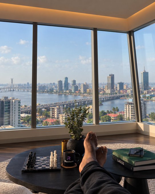 First-person view of feet propped on a dark coffee table with a chess set, plant, coffee mug, and books, looking out large windows at a city skyline and river with bridges.