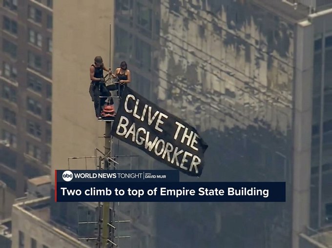 Two people sit atop the Empire State Building's antenna displaying a banner reading "CLIVE THE BAGWORKER," as reported on ABC World News Tonight with David Muir. The image connects to the caption about "coin pushin'" by referencing street performer slang, with "bagworker" being a derogatory term often used by carnival and street hustlers to refer to their patrons, adding irony to their high-profile stunt.