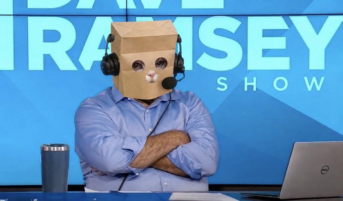 A man sits at a desk with arms crossed, wearing a paper bag over his head decorated with a cat face and a headset, in front of a "Dave Ramsey Show" backdrop featuring a Dell laptop and tumbler—visually embodying the absurd post's humor about blindly holding $ZCAT for "zividends" like a cat in a bag.