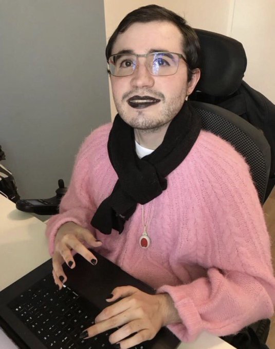 A digitally manipulated image of a man with glasses, dark lipstick, a black scarf, and a pink sweater sitting at a desk in front of a laptop.