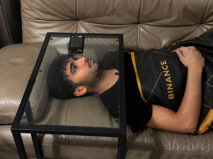 A man lies on a couch with a glass display case over his head holding a phone, while wearing a black shirt with "BINANCE" in gold lettering—humorously illustrating an extreme new way to check the Binance chart first thing in the morning.