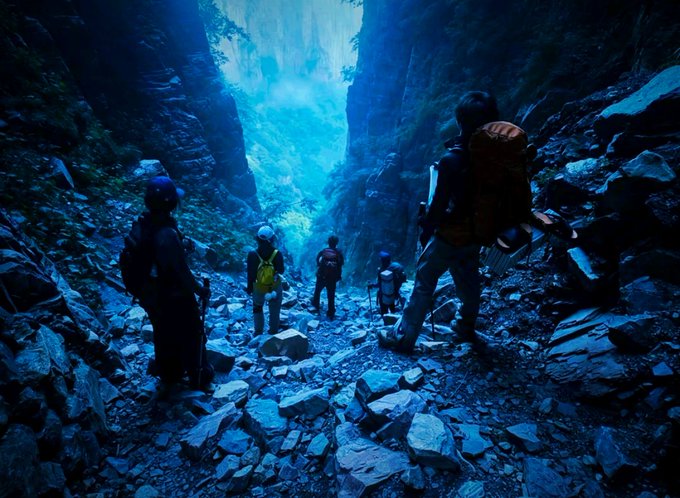 A group of hikers with backpacks exploring a rocky cave illuminated by ethereal blue light streaming in from above.