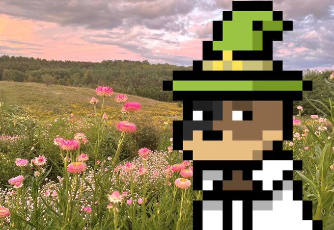 urn of pink flowers, a green hat with yellow trim, and a brown dog with black ears stand against a backdrop of trees and hills.