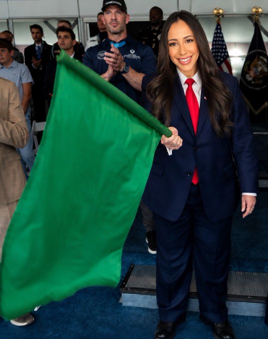 A woman in a navy suit and red tie waves a green flag in celebration, likely for a triumphant event, while onlookers including a man in a baseball cap applaud in the background near an American flag and an official seal. The image's celebratory green flag aligns with the post's triumphant message about Jackson Hole Day and successful investment gains.