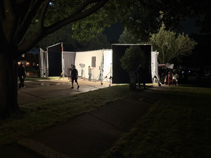 A nighttime behind-the-scenes photo of a film production set outdoors, featuring crew members, lighting equipment, and large diffusion panels under a tree.