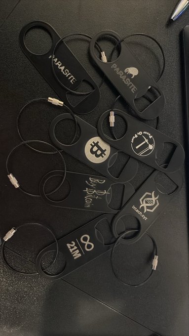 Several black paracord crypto-themed keychains are scattered on a dark surface, featuring logos and text for Bitcoin, 'Bitcoin to the Moon', IRA, 'Be Your Own Bank', and 'ZIM'.
