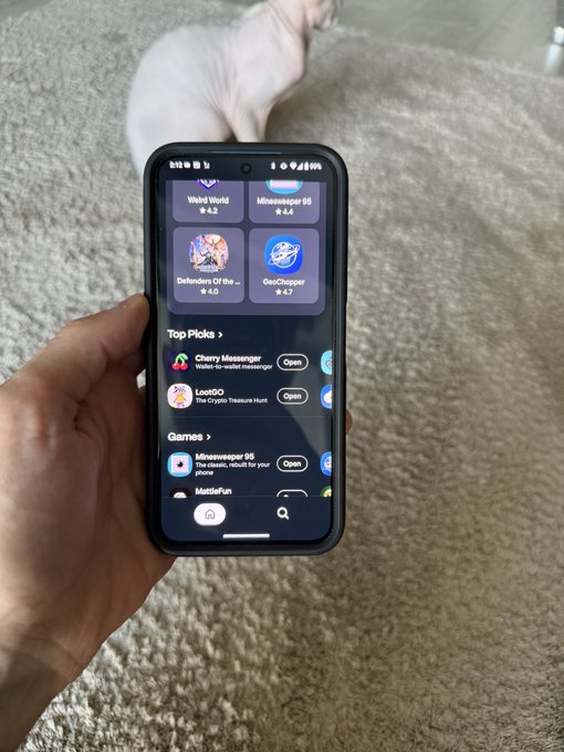 A hand holds a smartphone displaying an app store page for "MINESWEEPER 95" with a 4.4 rating, while a light-colored cat sits on a beige carpet in the background.