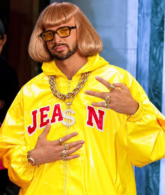 A man with a blonde bob haircut and yellow-tinted sunglasses wearing a yellow jacket with "JEAN" written across it, adorned with multiple gold chains and rings.