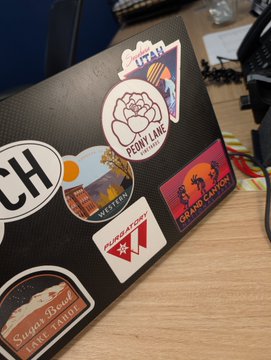 A close-up of a black textured laptop covered in various travel and location stickers, including 'Premium Lake Tahoe', 'Grand Canyon', 'Vermont', 'Hungary', and 'Sausalito'.