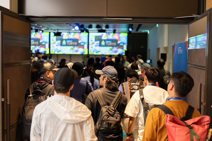 A crowd of people, many with backpacks and caps, walks through a doorway into a large hall with bright screens displaying colorful content.