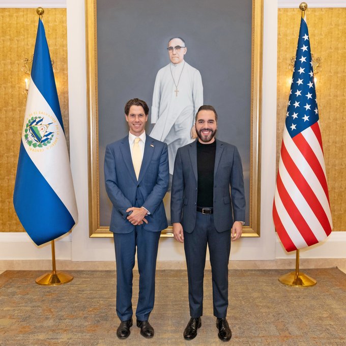 Two men in suits posing together in a formal setting, flanked by the flags of El Salvador and the United States, with a large portrait hanging behind them.