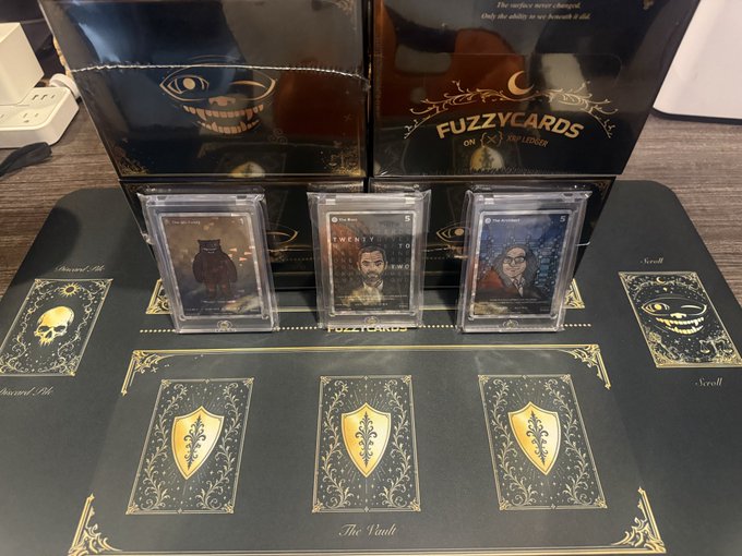 Three encased trading cards labeled "FUZZY CARDS" are displayed on "The Vault" playmat, with packaging visible in the background, celebrating the physical release of the collectible card series.