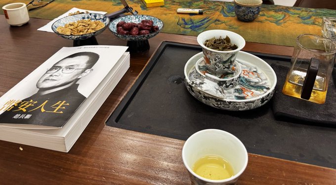 A Chinese tea ceremony setup sits on a dark wooden table, featuring an ornate gaiwan containing dark tea leaves, accompanied by a small tasting cup with golden liquor, dried dates and nuts in decorative dishes, and a book titled "腾安人生" (Teng An's Life) with the subtitle "战术篇" (Tactical Edition). The contemplative tea-drinking scene visually reinforces the post's message about mastering emotional discipline in market timing—suggesting that proper investment decisions, like the patient ritual of tea preparation, require calm reflection rather than panic-driven impulses at market extremes.