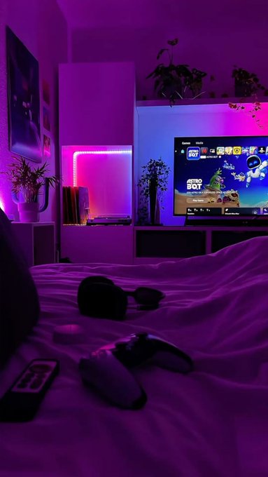 A dimly lit bedroom illuminated by purple and pink LED lights, featuring a gaming console and a TV displaying a game menu.