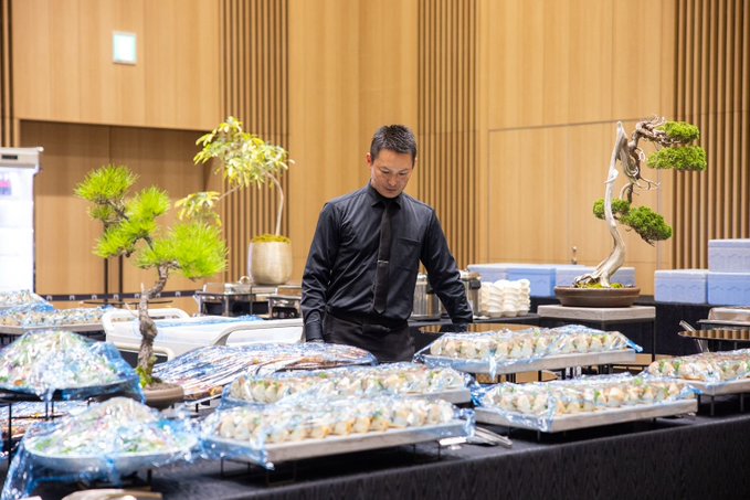 A sushi chef in a black uniform and tie standing behind a buffet table filled with trays of sushi, with decorative bonsai trees and a minimalist wooden interior in the background.