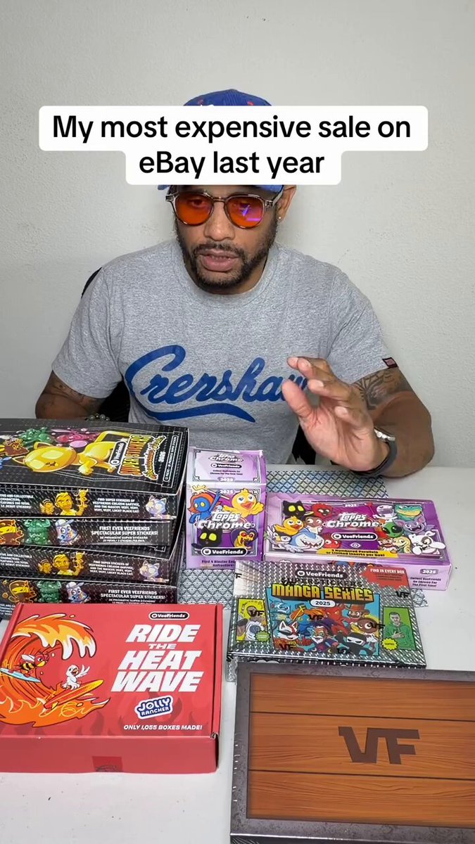The image features a man sitting at a table with numerous boxes of toys surrounding him, including a game box. He appears to be taking a break from sorting these items or possibly selling them online. The text in the photo indicates that he has sold 7,223 items last year and is actively seeking out new VeeFriend trading cards to trade for more of his own goods. He is specifically looking for bangers to complete his collection, as indicated by the term "trade".