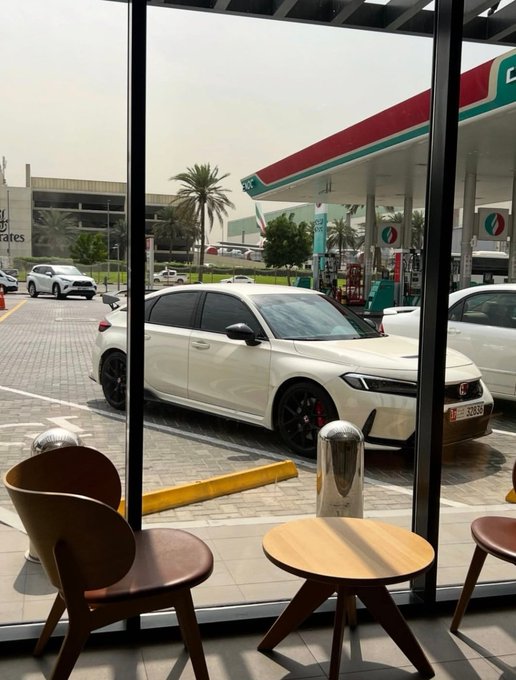 A white Honda Civic Type R is parked at an ENOC gas station, viewed from inside a café with wooden chairs and a small table in the foreground.