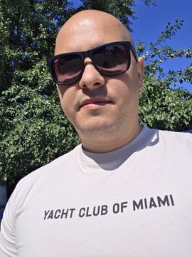 A bald man wearing sunglasses and a white long-sleeve shirt with 'YACHT CLUB OF MIAMI' printed on the front, posing outdoors with green trees and blue sky in the background.