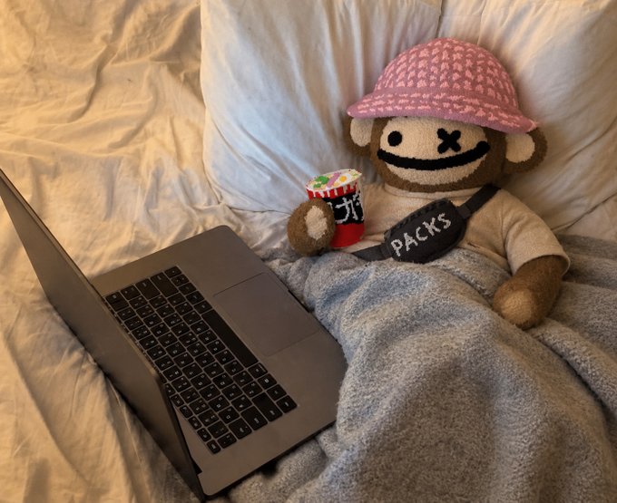 A stuffed monkey toy wearing a pink patterned bucket hat and a 'PACKS' sash lies in bed under a gray blanket next to an open laptop, holding a cup of instant noodles.