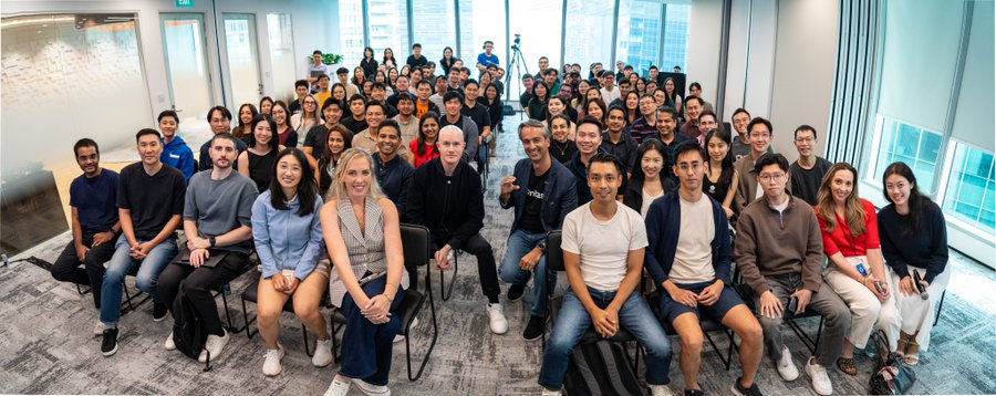 A large group photo of a diverse team gathered in a modern office space with floor-to-ceiling windows, seated and standing in rows for a group picture.