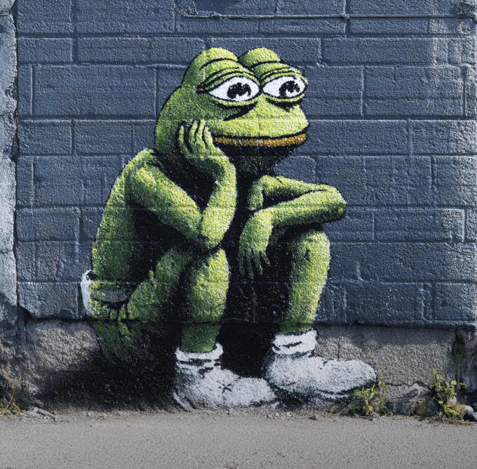 A graffiti of a green frog wearing white sneakers sits hunched over on a dark brick wall.