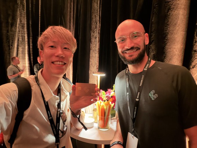 Two men wearing Token2049 lanyards smile at the camera at an event, with one holding a small golden or red object between them near a small table with flowers and a lamp.