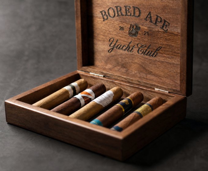 The image features a wooden cigar box with five different colored cigars inside, including a white cigar, an orange cigar, a brown cigar, a blue cigar, and a yellow cigar. The box has a label that reads "Bored Ape Yachty Club", indicating it is a collector's item from the Bored Ape Yachty Club.